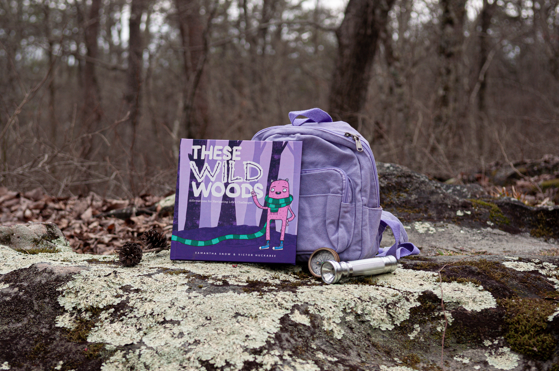 These Wild Woods book with a lavender backpack, compass, and flashlight in the woods